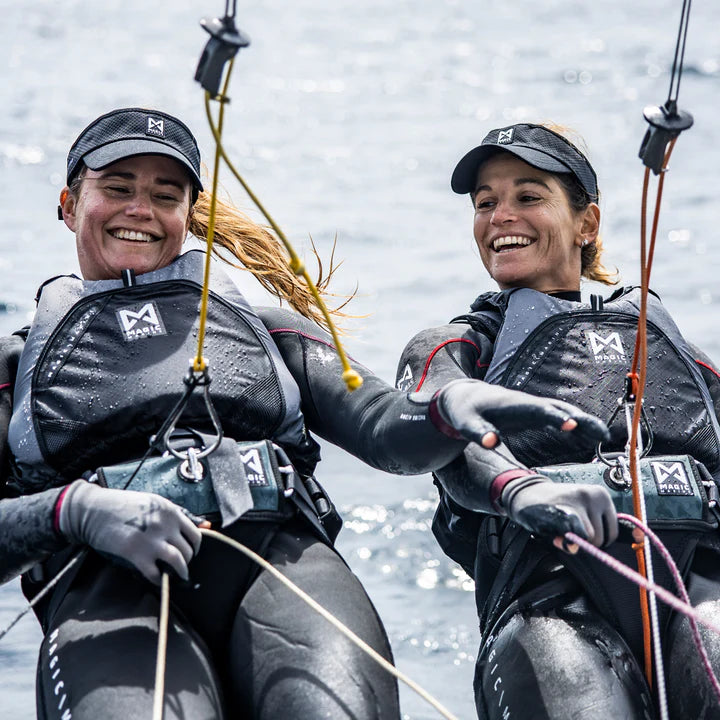 Two smiling women in wetsuits and caps on a sailboat with a water background.